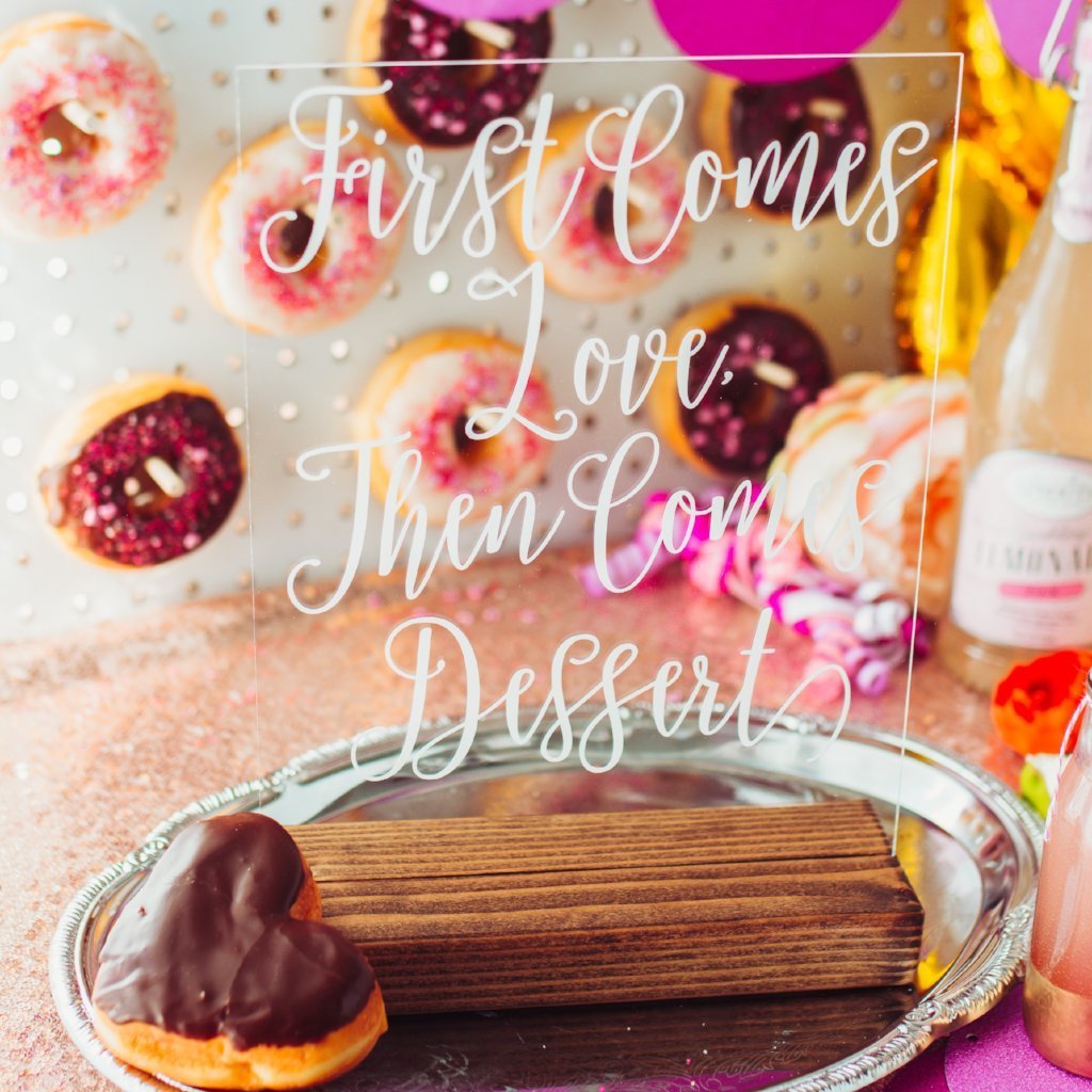 Clear acrylic dessert sign that reads "first comes love, then comes dessert" in white with a pink and chocolate donut wall backdrop.